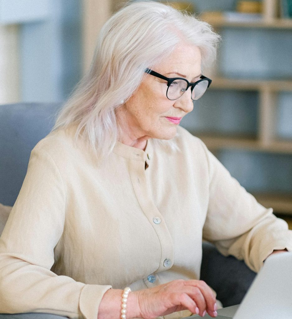 Elderly woman with glasses typing on a laptop in a cozy home setting, embodying remote work lifestyle.